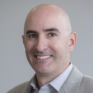 A bald man smiling at the camera wearing a grey suit jacket and grey button up shirt