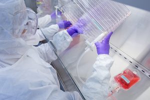 A person in a white lab coat and goggles holding equipment under a hood