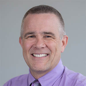 A man with salt and pepper hair smiling at the camera wearing a purple button up shirt.
