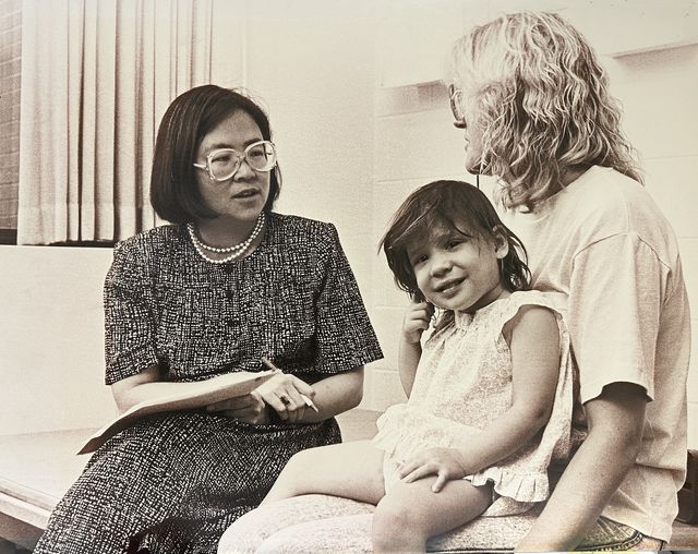 A woman holding a clipboard speaking to a woman with a young child on her lap