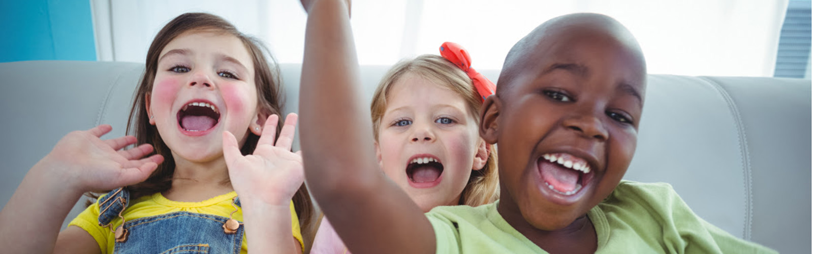 Three young children sitting on white leather couch smiling with mouths open. Front child is a black boy wearing a green shirt. Behind him is a white girl with blonde hair wearing a red bow. Next to her is a white girl with brown hair wearing overalls and holding her hands up towards the sky.