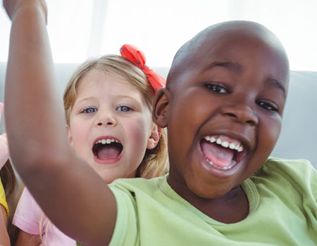 two children smiling with mouths open. Front child is a black boy wearing a green shirt. Behind him is a white girl with blonde hair wearing a red bow.