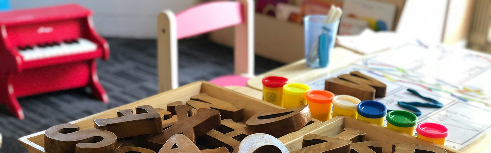 Picture of preschool table with wood letters, multi-color play dough canisters on it and red miniature piano and book shelf in background