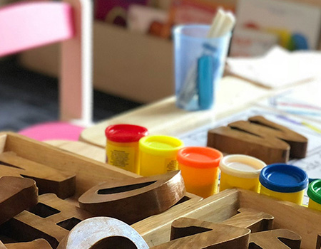 Picture of preschool table with wood letters, multi-color play dough canisters on it. Bookshelf in the background.