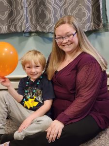 A young blonde boy holding an orange balloon sitting on the floor with his mother