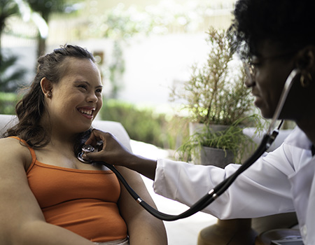 black female doctor with glasses using a stethoscope on a white young adult with down syndrome
