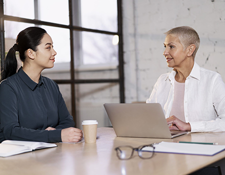 Two women sitting at a conference table. The younger woman is Asian with a dark collared shirt and long black hair in a ponytail. The other woman has short, light colored hair and is wearing a pink shirt and white collared shirt over it.