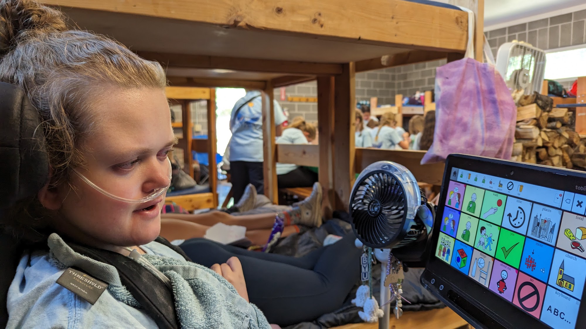 Young white girl using AAC device at camp surrounded by bunk beds and other campers.