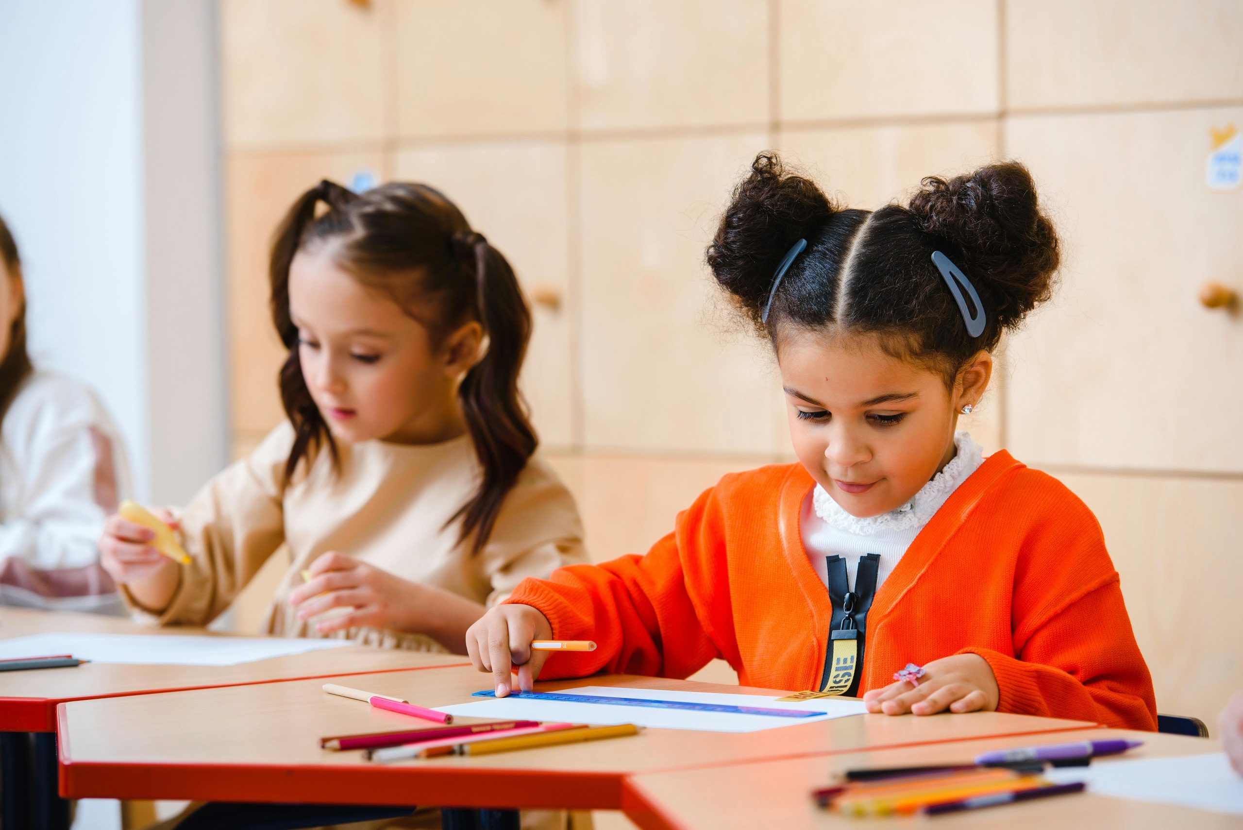 Two little girls sitting at a desk coloring/writing. One girl is white with dark brown hair in pig tails. She is wearing a cream colored top. The other girl has dark space buns with barrettes in her hair. She is wearing an orange cardigan and a white shirt under it.