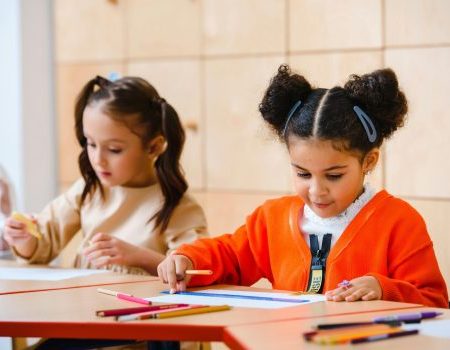 Two little girls sitting at a desk coloring/writing. One girl is white with dark brown hair in pig tails. She is wearing a cream colored top. The other girl has dark space buns with barrettes in her hair. She is wearing an orange cardigan and a white shirt under it.