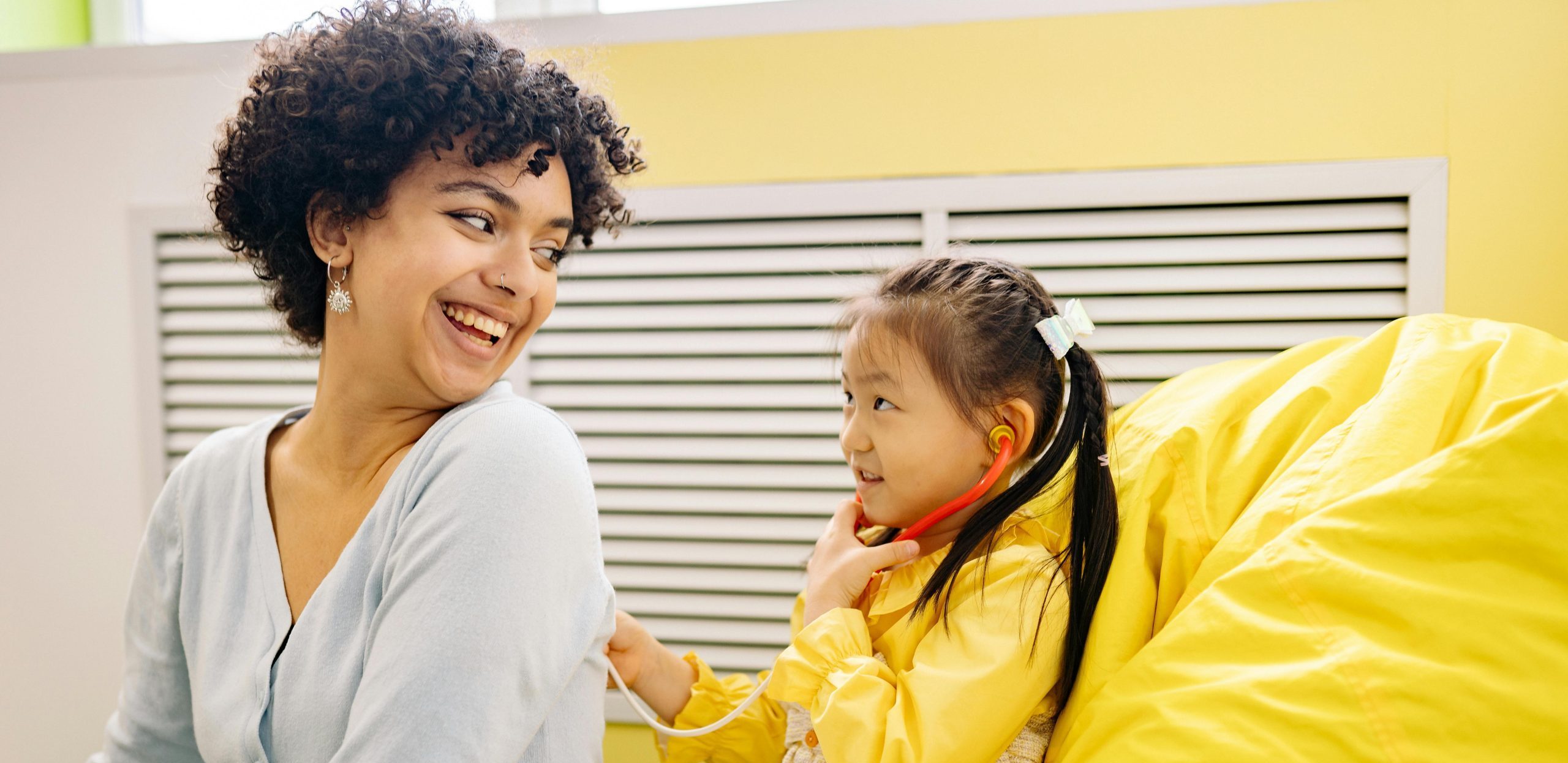 Young Asian girl in a yellow top with dark hair braided behind her with a white bow is using a toy stethoscope on an adult's back. The adult is female and is wearing a light blue top. She has brown curly hair and is smiling at the child.