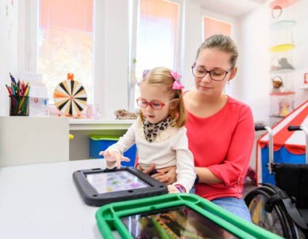 Little girl sitting on adult's lap in clinic space using AAC device. Little girl a blonde white child, wearing a white shirt, cheetah print scarf, red glasses, and pink and purple bows in her hair. Adult is a white female wearing a pink top and jeans. A wheelchair is pictured in the background.