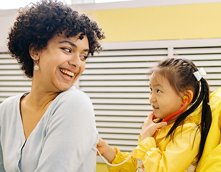 Young Asian girl in a yellow top with dark hair braided behind her with a white bow is using a toy stethoscope on an adult's back. The adult is female and is wearing a light blue top. She has brown curly hair and is smiling at the child.