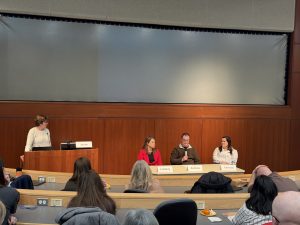 Dr. Boyette Anderson stands in front of the podium. Next to her, the three in-person panelist are sitting at a table. Higgins holds the microphone.