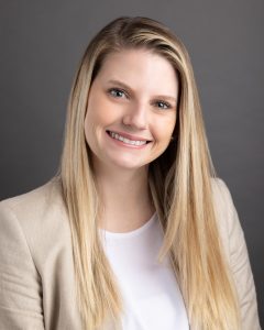 Woman with blonde hair wearing a tan jacket and white shirt smiling at the camera