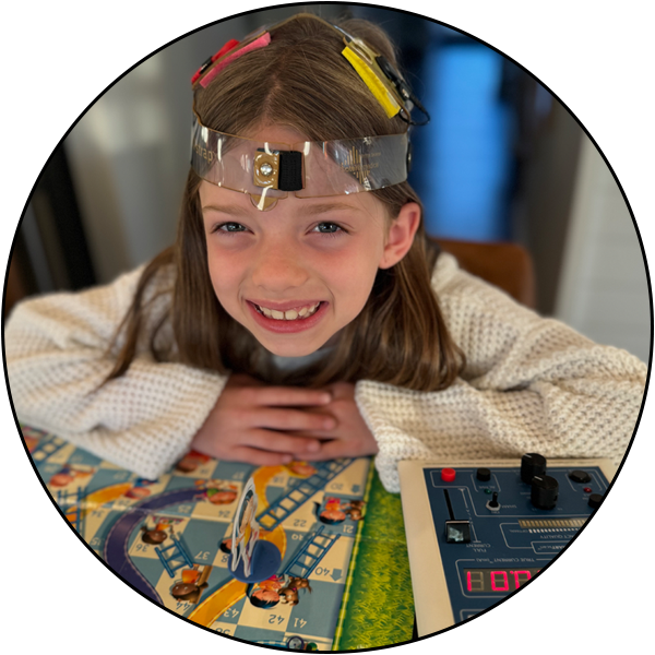 A smiling girl sits at a table with a tDCS cap on her head