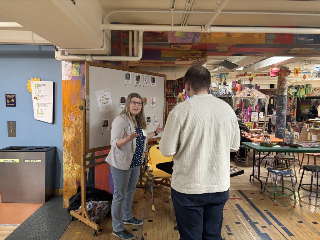 Shutts stands in front of a whiteboard with her arms open as she talks to a man who has his back to the camera.