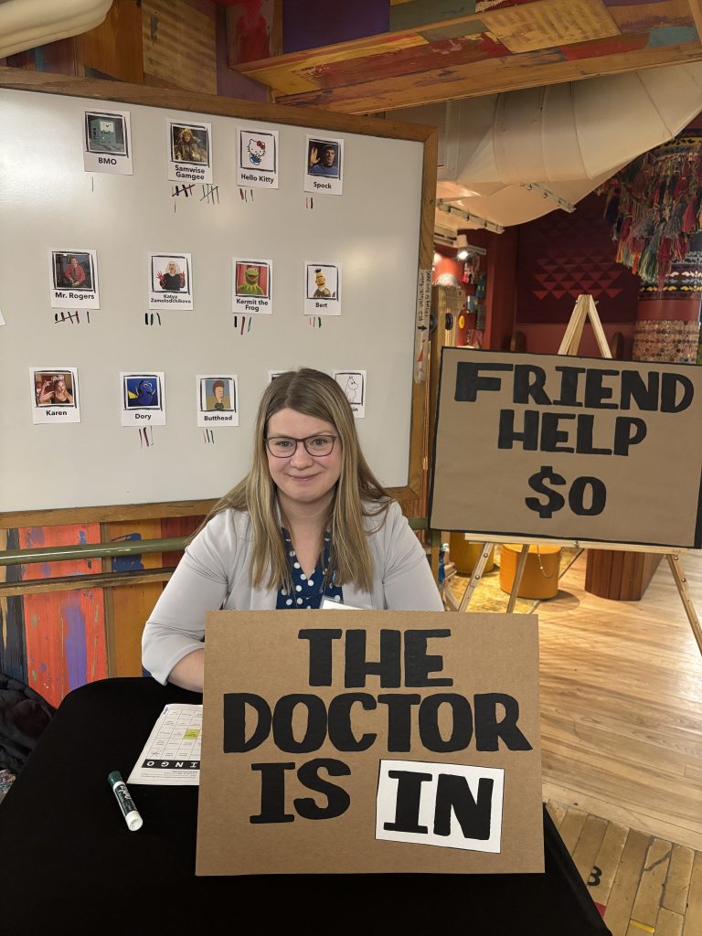 Shutts, with medium length blond hair and glasses, wearing a grar blazer over a blue shirt sits behind a carton sign that reads the doctor is in. Behind her is a sign that reads friend help $0.