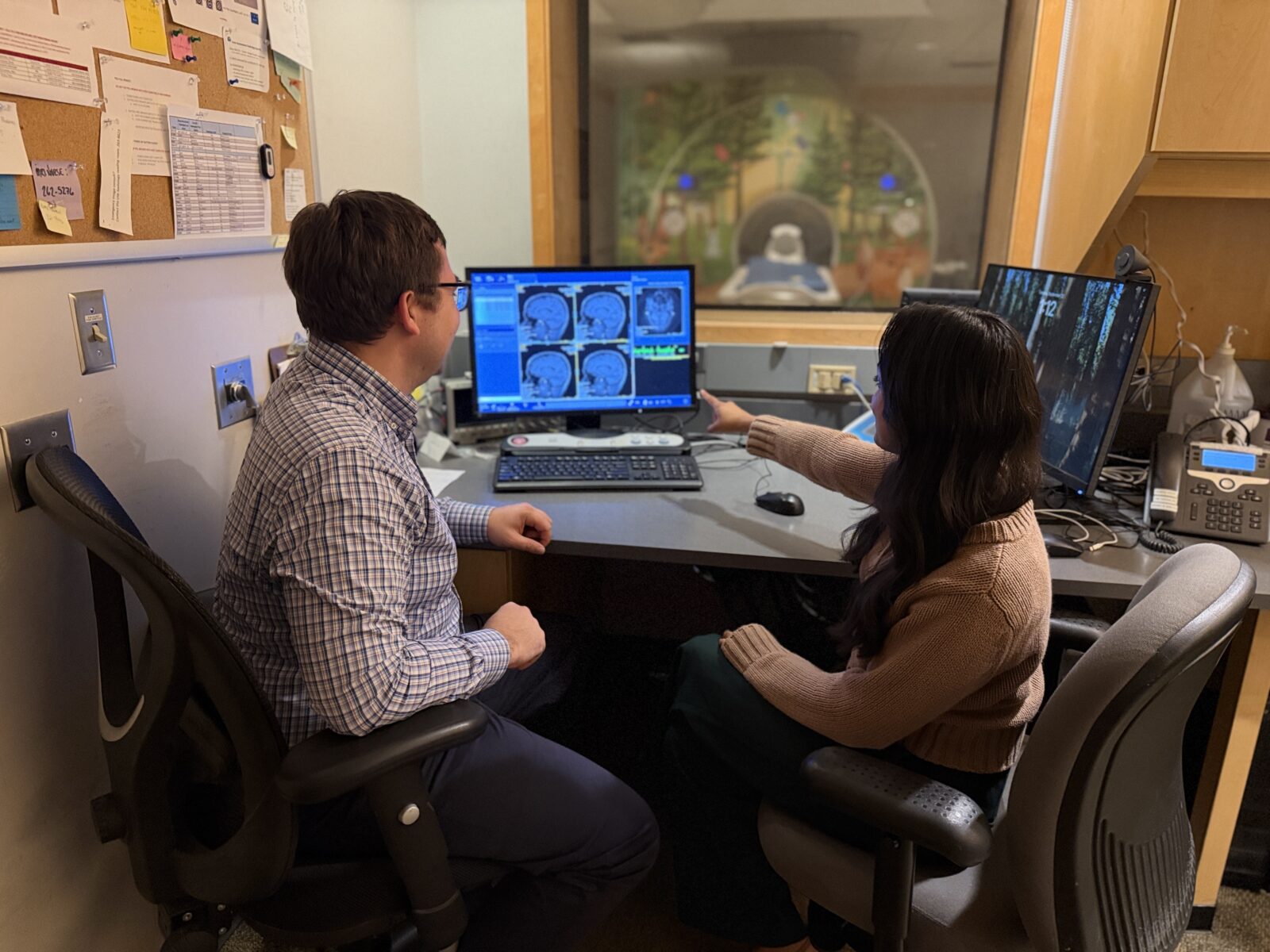 Doug and Leela sitting by a computer, with Leela pointing to brain MRI images in the screen. MRI scanner is in the back.