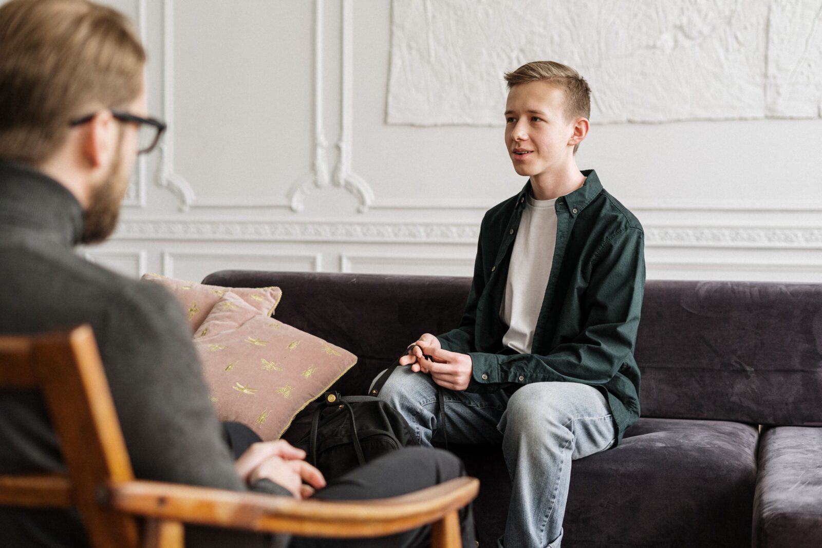 Teenager sitting on couch wearing a light grey t shirt and green button down holding a black backpack, talking to adult therapist wearing a grey sweater and black pants.