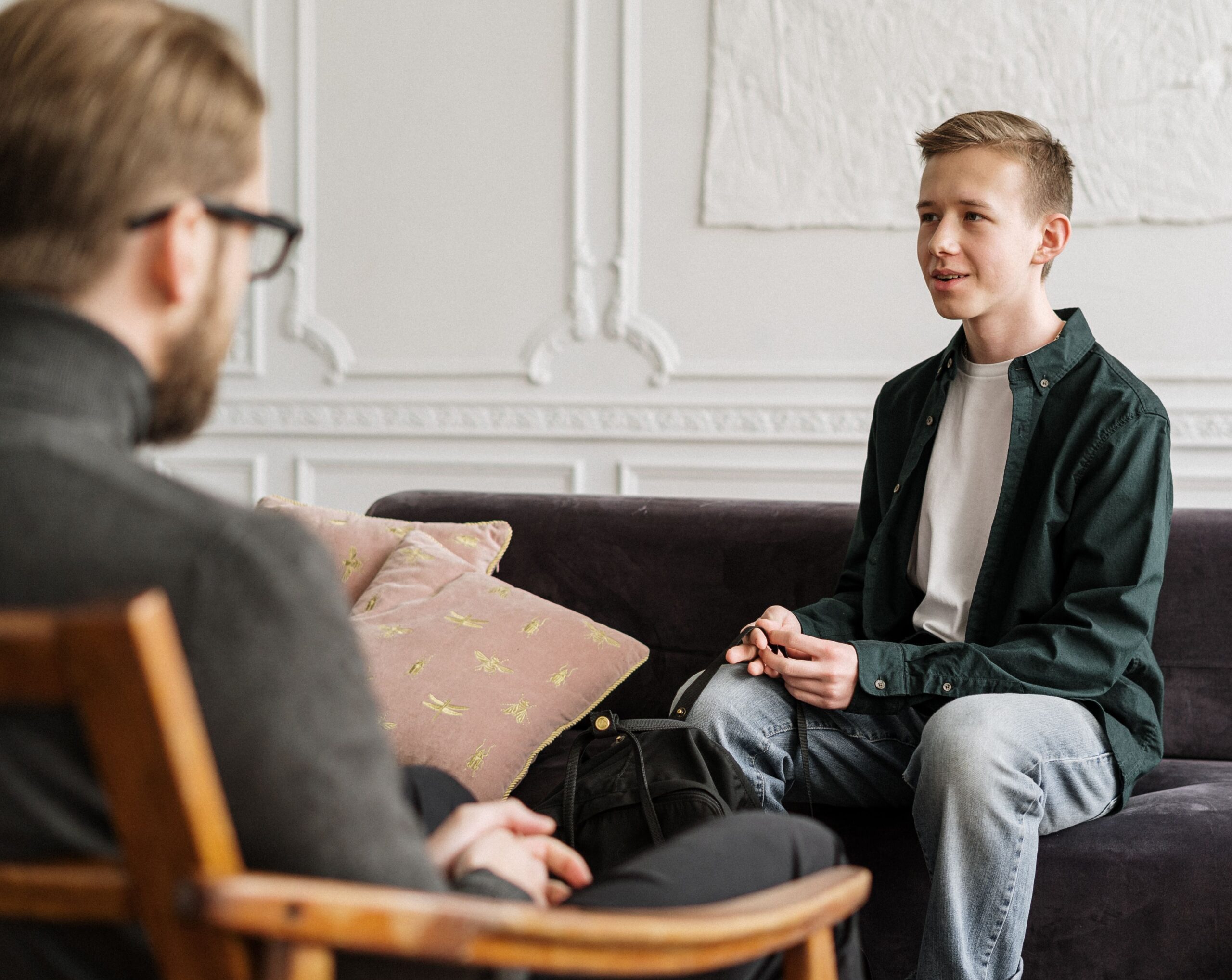 Teenager sitting on couch wearing a light grey t shirt and green button down holding a black backpack, talking to adult therapist wearing a grey sweater and black pants.
