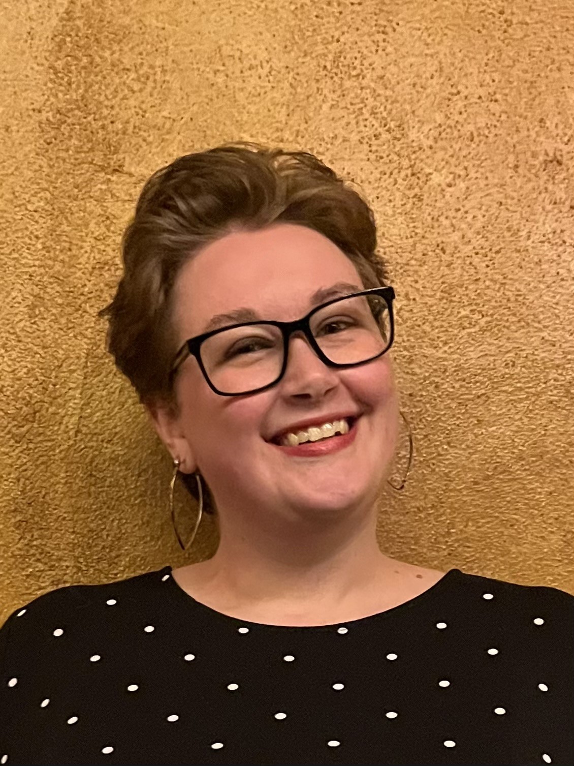  Helen, a smiling white person with short brown hair and glasses wearing a black and white polka dot dress and silver hoop earrings.